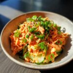 A close-up of a vibrant orange spicy kani salad served in a light brown bowl, topped with fresh green onions and sesame seeds.