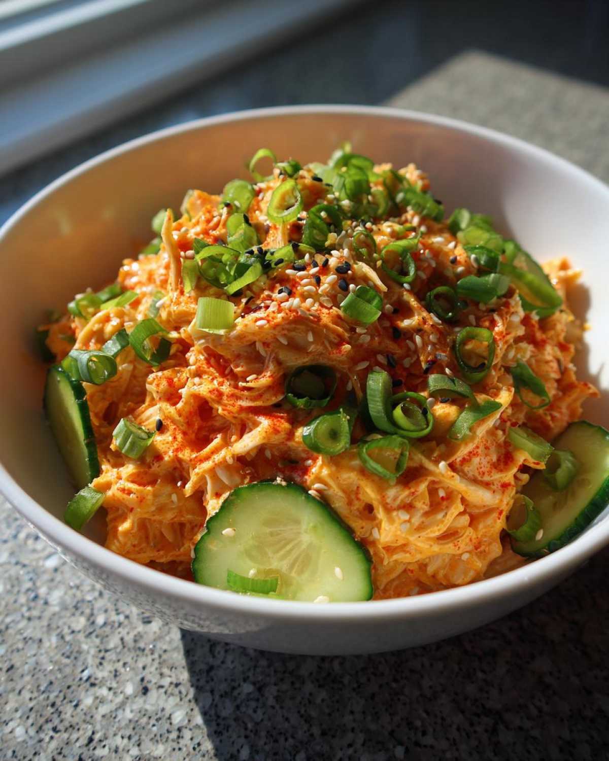 A close-up of a vibrant, creamy spicy kani salad topped with green onions and sesame seeds, garnished with cucumber slices.