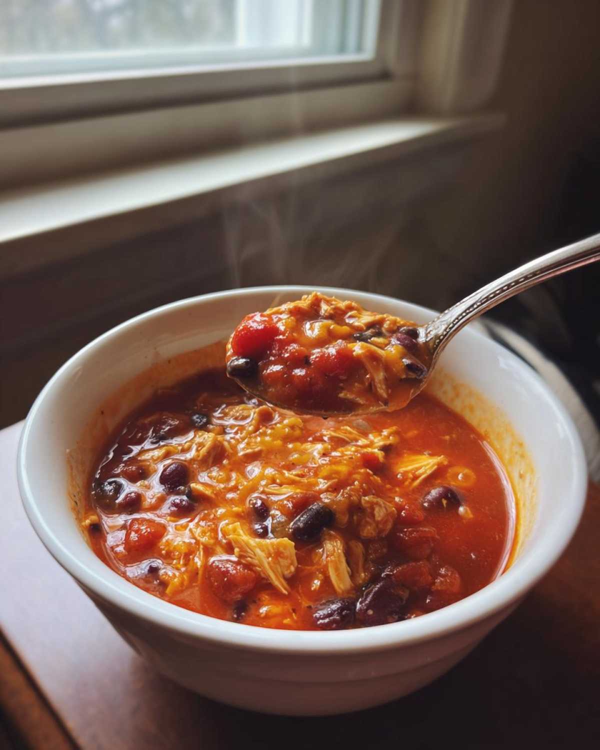 A spoonful of steaming hot slow cooker buffalo chicken chili, rich in shredded chicken and black beans, being lifted from a white bowl.