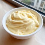 A close-up of thick, pale yellow homemade mayonnaise swirled beautifully in a small white bowl.