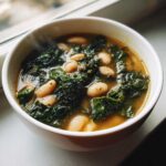 Close-up of a steaming white bowl filled with Tuscan white bean soup, featuring white beans and dark green kale in a savory broth.