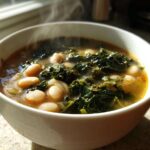 A steaming white bowl filled with hearty Tuscan white bean soup featuring cannellini beans and dark leafy greens.
