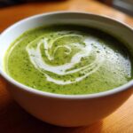 Close-up of a bowl of bright green zucchini soup topped with a swirl of cream and herbs.