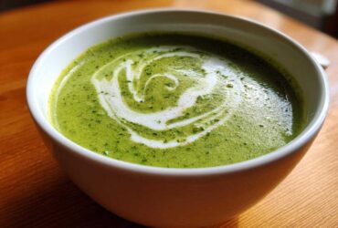 Close-up of a bowl of bright green zucchini soup topped with a swirl of cream and herbs.
