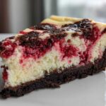 Close-up of a rich slice of white chocolate raspberry cheesecake showing layers of dark crust, creamy filling, and bright red raspberries.