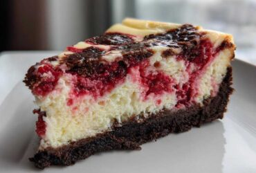 Close-up of a rich slice of white chocolate raspberry cheesecake showing layers of dark crust, creamy filling, and bright red raspberries.