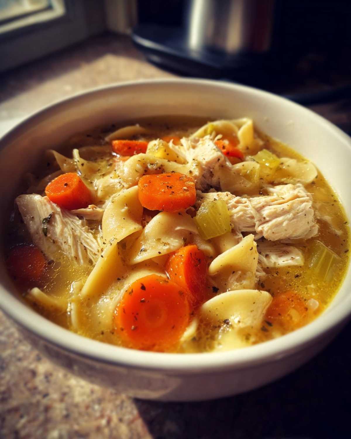 Close-up of a white bowl filled with rich chicken noodle soup, featuring shredded chicken, egg noodles, carrots, and celery.