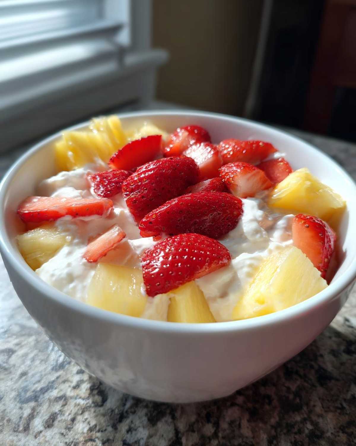 A white bowl filled with creamy fruit dip topped with fresh sliced strawberries and pineapple chunks.