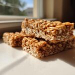Three stacked, chewy, homemade protein bars made with oats, sitting on a white surface in bright sunlight.