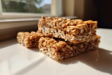 Three stacked, chewy, homemade protein bars made with oats, sitting on a white surface in bright sunlight.