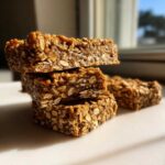 Stack of three chewy, homemade protein bars made with visible rolled oats, set against a bright window.
