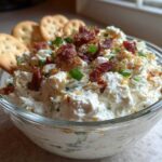 A close-up of creamy Million Dollar Dip topped with bacon bits and green onions, served with crackers.