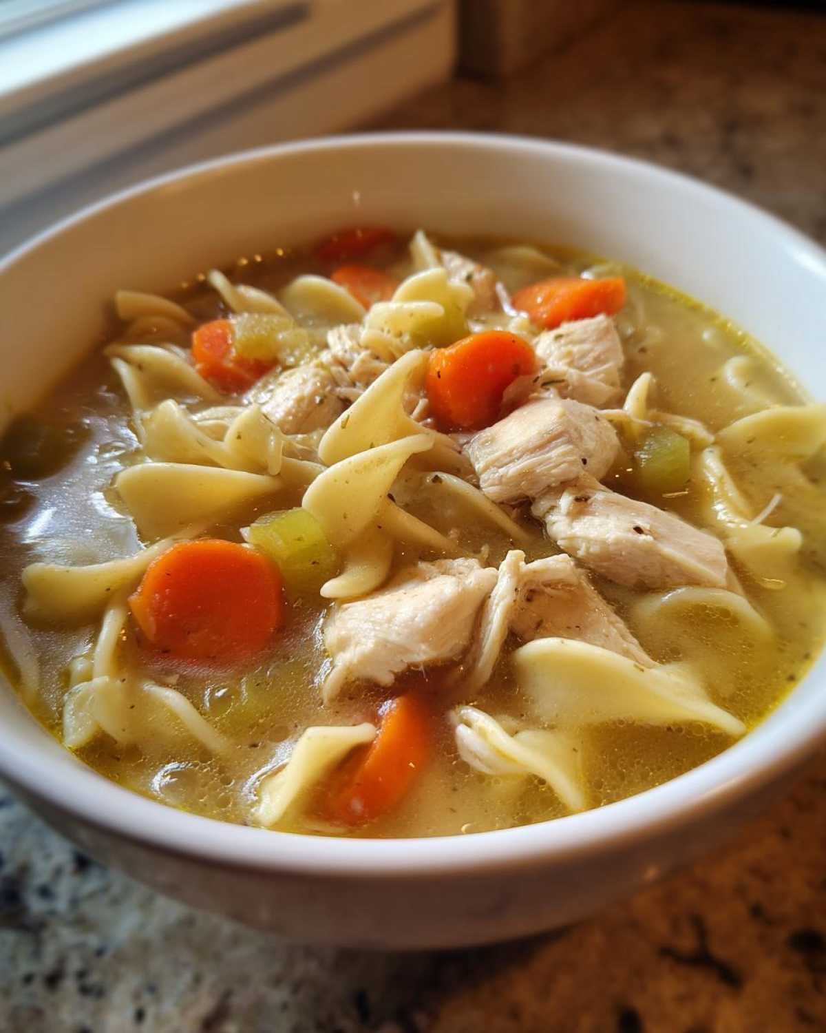 Close-up of a white bowl filled with amazing 1-pot chicken noodle soup, showing broth, egg noodles, chunks of chicken, carrots, and celery.