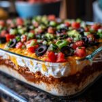 Close-up of a delicious 7 layer dip in a glass baking dish, topped with olives, tomatoes, and avocado.