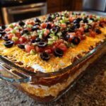 Close-up of a vibrant, fully assembled 7 layer dip in a clear glass dish, topped with cheese, olives, tomatoes, and green onions.