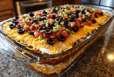 Close-up of a vibrant, fully assembled 7 layer dip in a clear glass dish, topped with cheese, olives, tomatoes, and green onions.