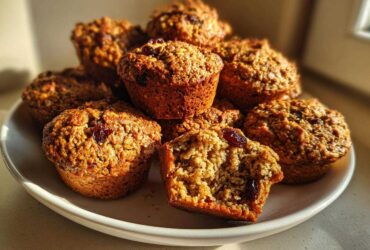 A stack of freshly baked, golden brown bran muffins with visible oats and dried cranberries, one muffin is broken open.