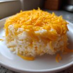 A molded serving of creamy white rice generously topped with bright orange shredded cheddar cheese, ready to eat as cheesy rice.