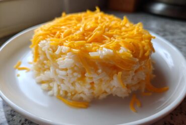 A molded serving of creamy white rice generously topped with bright orange shredded cheddar cheese, ready to eat as cheesy rice.