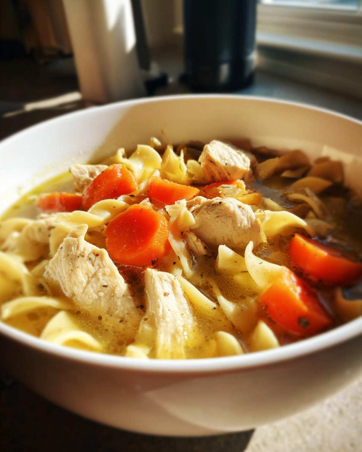 Close-up of a white bowl filled with homemade chicken noodle soup, featuring chunks of chicken, egg noodles, and bright orange carrots.