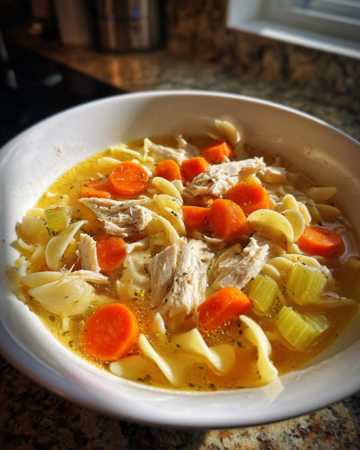 Close-up of a white bowl filled with amazing chicken noodle soup, featuring shredded chicken, carrots, celery, and egg noodles.