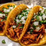 Close-up of three street-style chicken tacos filled with shredded meat, topped with diced white onion and fresh cilantro.