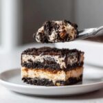 A fork lifts a bite-sized piece of layered oreo dirt cake above a square serving on a white plate.