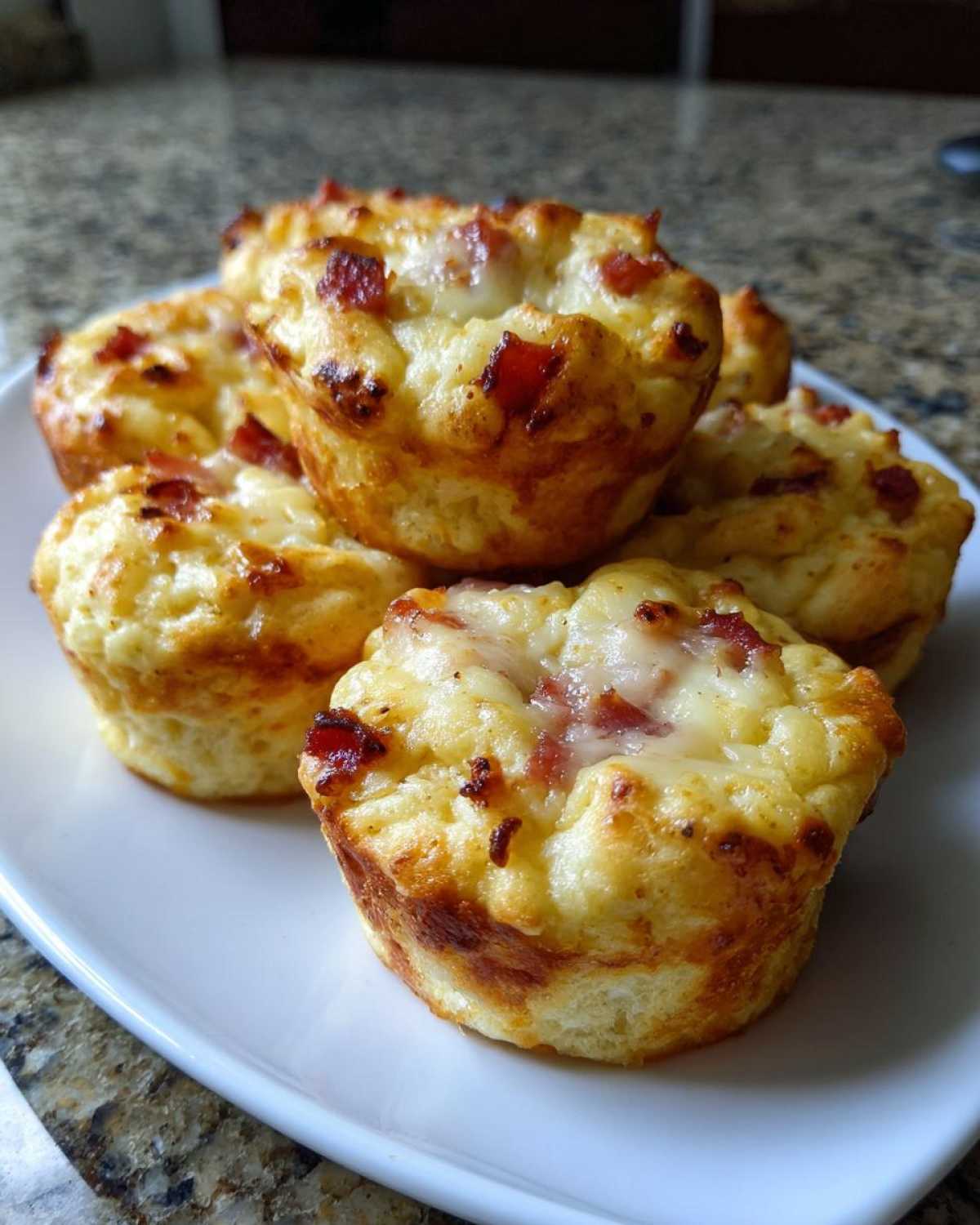 A stack of freshly baked, golden brown pizza muffins topped with melted cheese and bits of meat on a white plate.