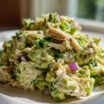 A close-up, bright photo of creamy avocado chicken salad featuring shredded chicken, chunks of avocado, and diced red onion.