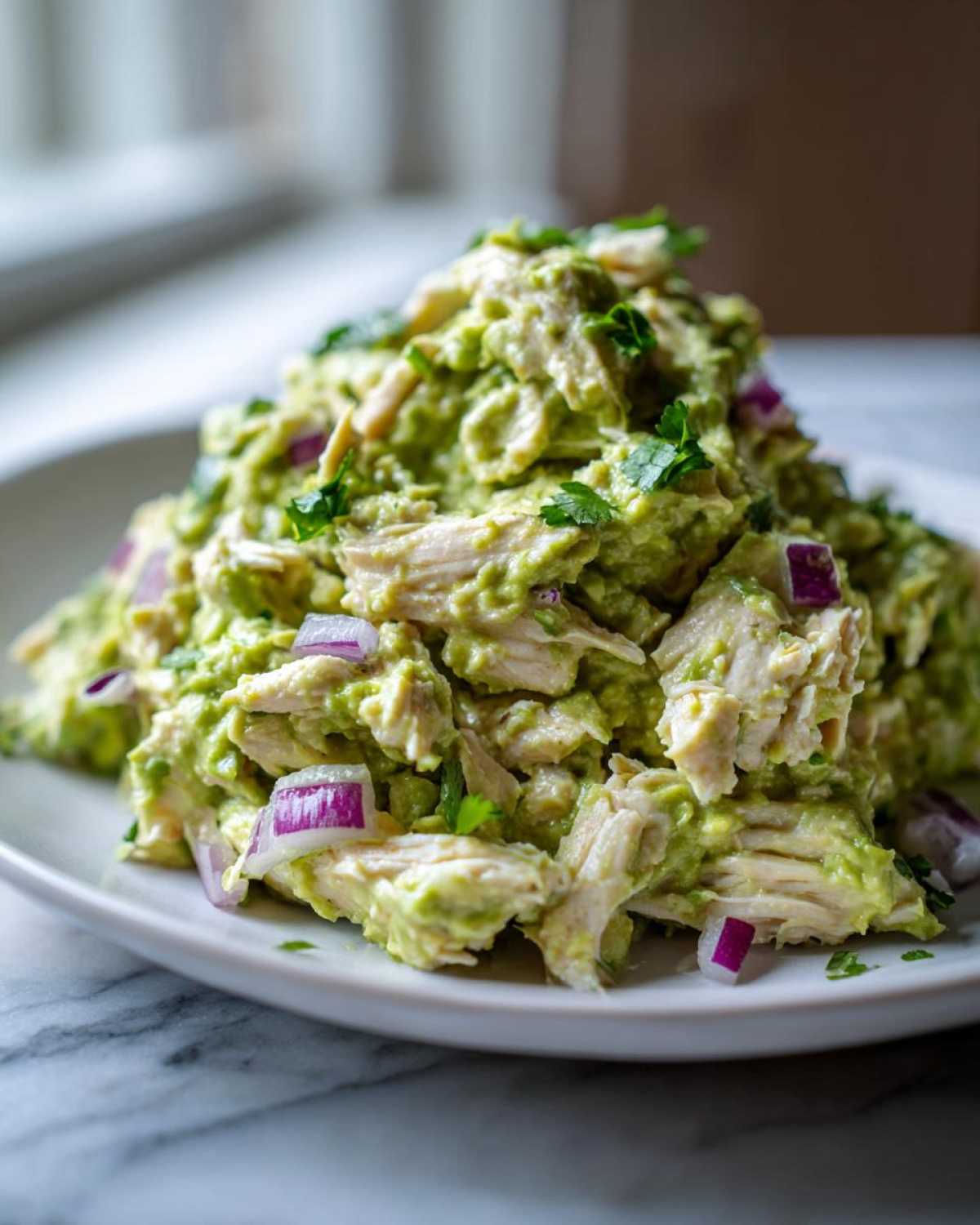 A mound of creamy avocado chicken salad mixed with shredded chicken and diced red onion, garnished with parsley.