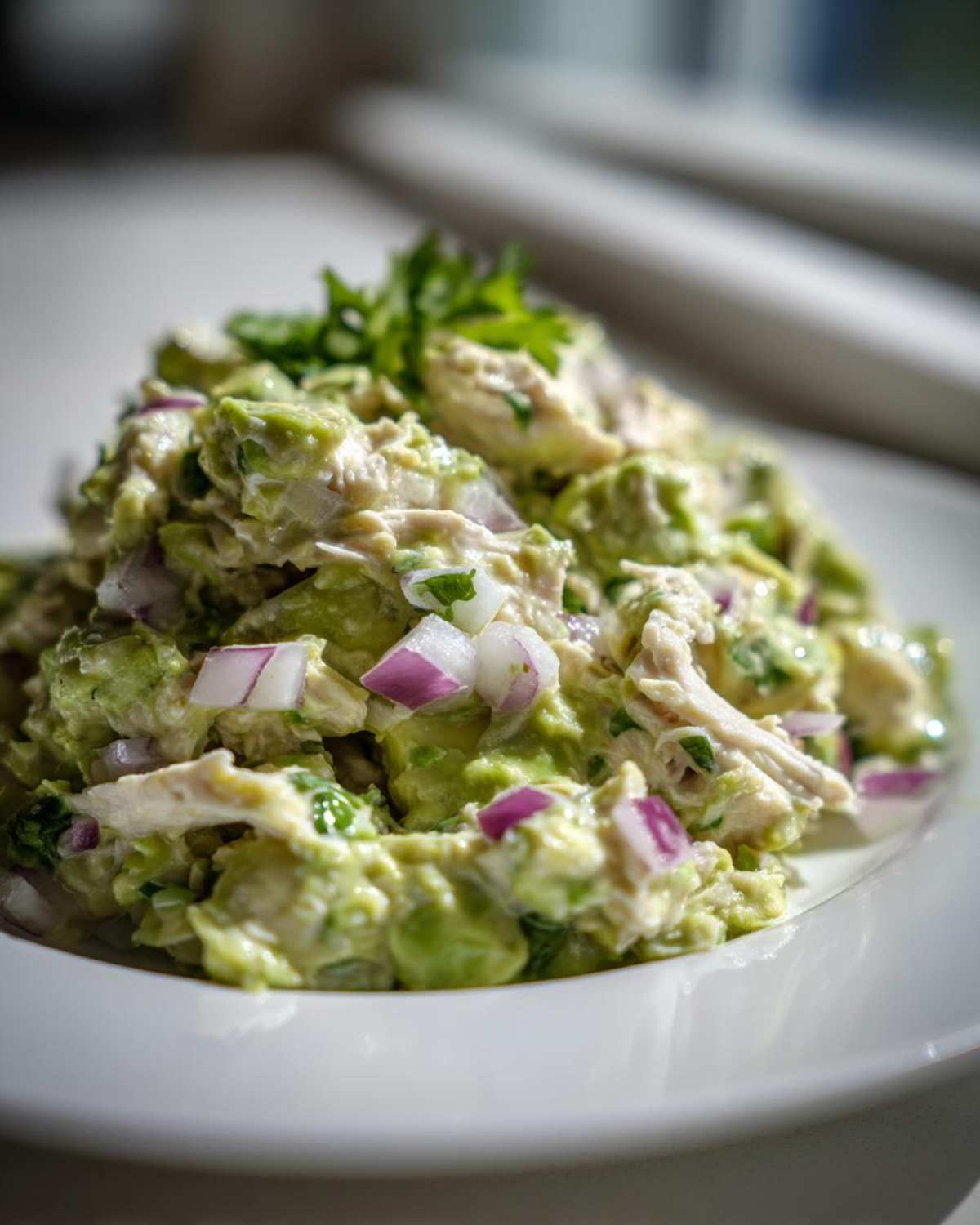A close-up, bright photo of creamy avocado chicken salad mixed with red onion chunks, served on a white plate.