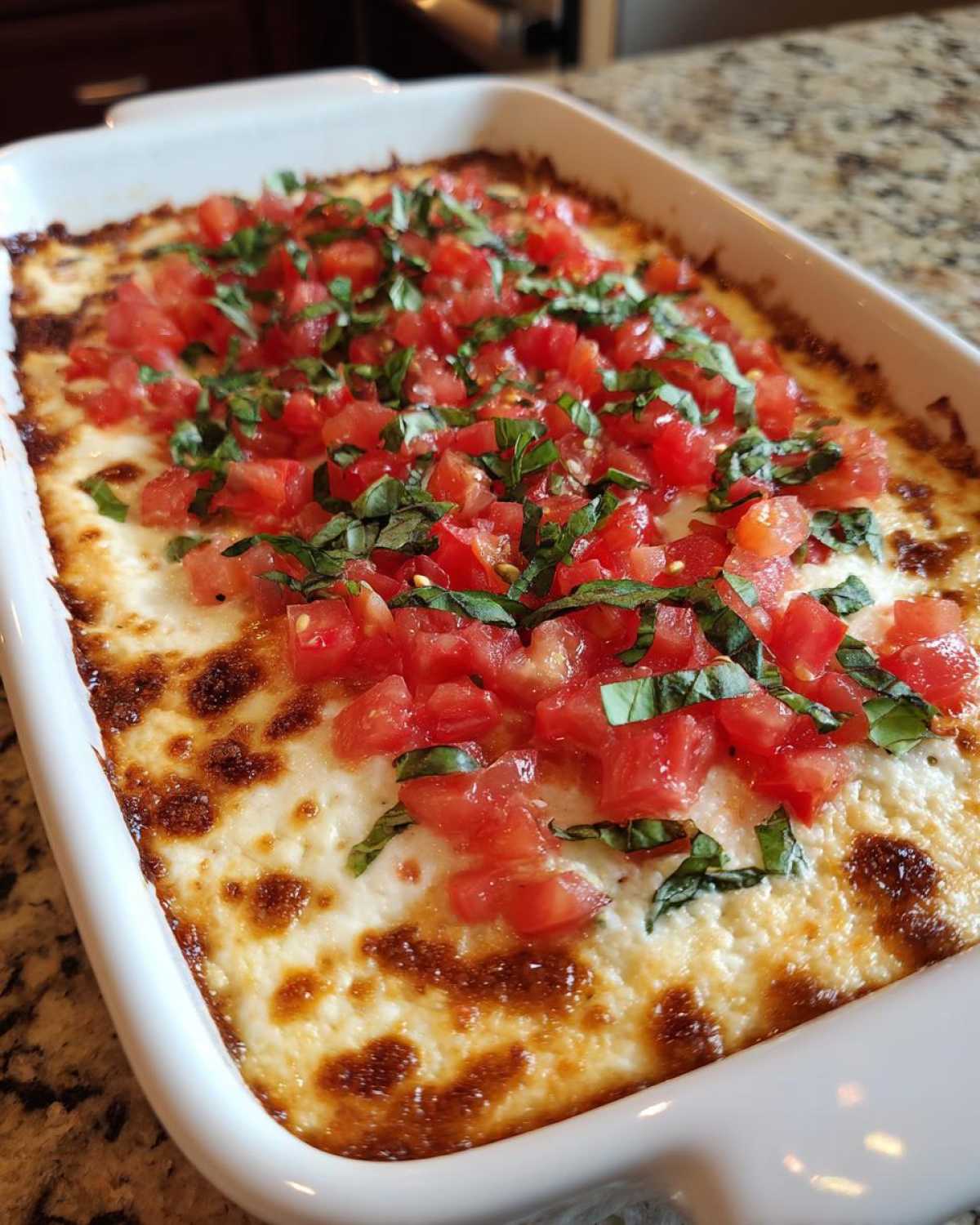 Close-up of a baked 3-cheese bruschetta dip topped with fresh diced tomatoes and basil in a white rectangular dish.