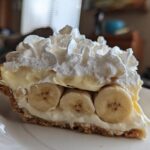 A close-up of a thick slice of banana cream pie recipe showing layers of graham cracker crust, banana slices, custard, and whipped cream.