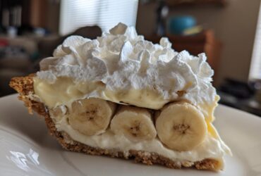 A close-up of a thick slice of banana cream pie recipe showing layers of graham cracker crust, banana slices, custard, and whipped cream.