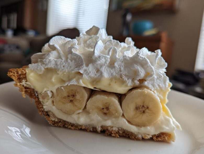 A close-up of a thick slice of banana cream pie recipe showing layers of graham cracker crust, banana slices, custard, and whipped cream.