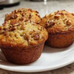 Three freshly baked banana nut muffins with a crunchy pecan topping served on a white plate.