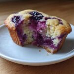 A close-up of a blueberry protein muffins with a bite taken out, showing the moist, purple-streaked interior.