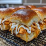 Close-up of several glossy-topped buffalo chicken sliders filled with shredded orange chicken and drizzled with white ranch sauce.