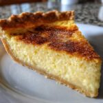 A close-up slice of rich buttermilk pie showing a creamy yellow filling and a beautifully caramelized, browned top.