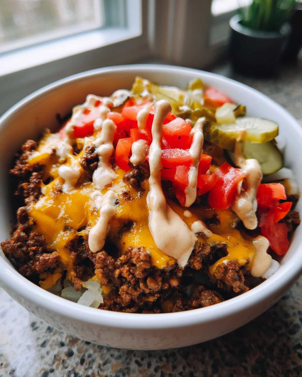 A close-up of a white bowl filled with a cheeseburger bowl featuring seasoned ground beef, melted cheddar cheese, diced tomatoes, pickles, and a drizzle of special sauce.