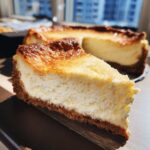 Close-up of a thick slice of baked cheesecake recipe showing a creamy white filling and dark graham cracker crust.