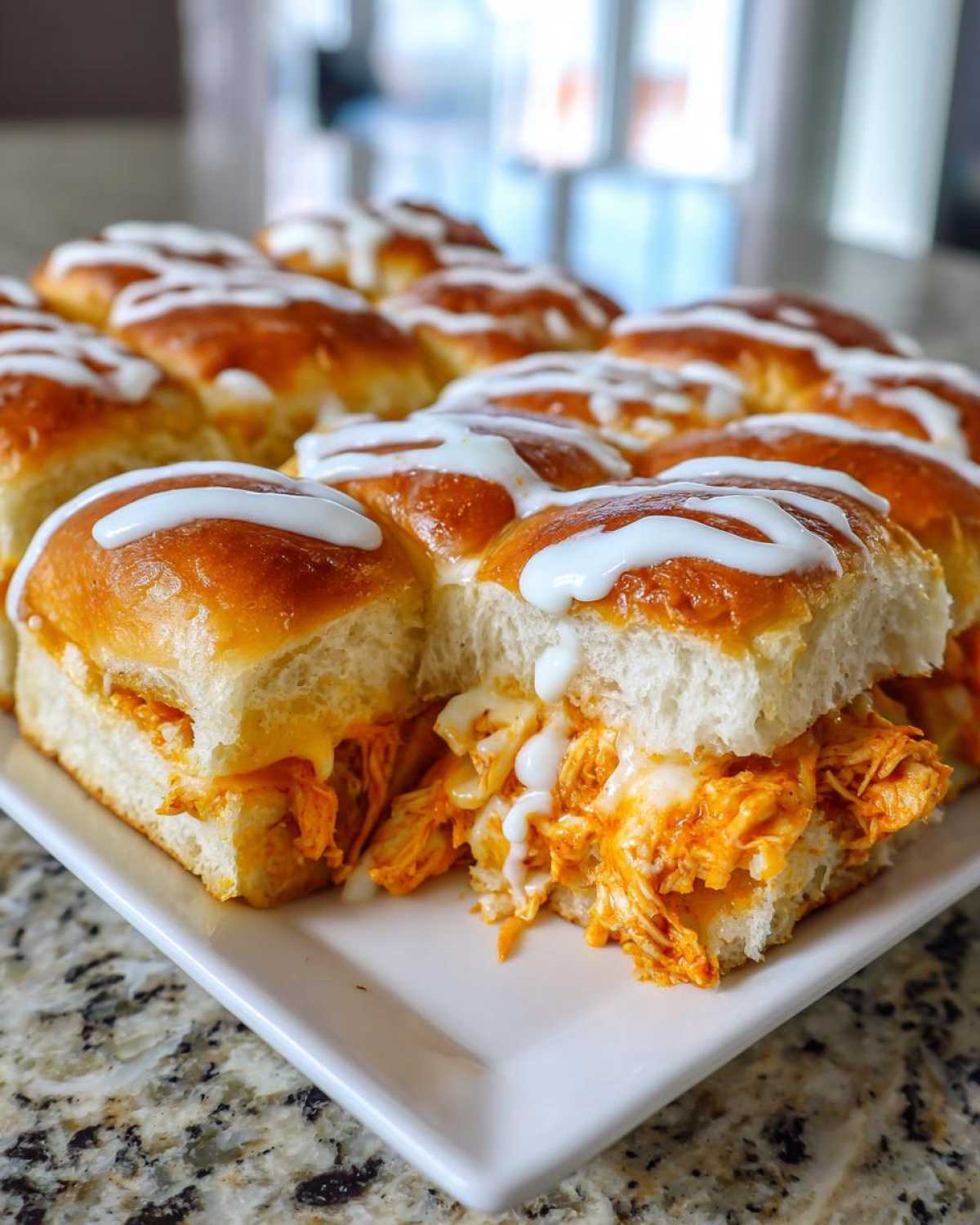 A platter of freshly baked buffalo chicken sliders, filled with shredded orange chicken and cheese, drizzled with white sauce.