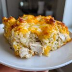 A close-up of a square serving of cheesy chicken and rice, topped with melted, browned cheddar cheese.