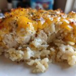 A close-up serving of baked cheesy chicken and rice, topped with melted, slightly browned cheddar cheese and herbs.