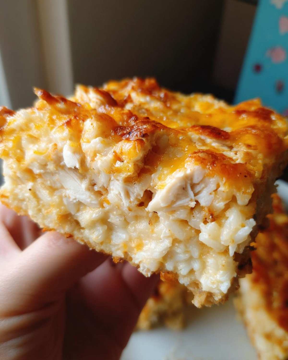 A hand holding a thick, square slice of cheesy chicken and rice casserole with a golden-brown, bubbly cheese topping.