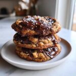 A stack of three thick, chewy chocolate chip cookies topped with melted chocolate and flaky sea salt.
