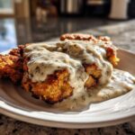 Close-up of golden brown chicken fried steak smothered in creamy, herbed white gravy on a white plate.