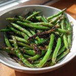 A close-up of bright green, glossy chinese green beans with garlic sauce and charred bits in a white bowl.