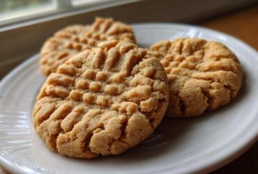 Close-up of three perfectly baked peanut butter cookies featuring the classic crisscross fork marks, resting on a white plate.