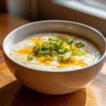 A close-up of a bowl of creamy cauliflower soup topped with melted cheese drizzle and fresh green scallions.